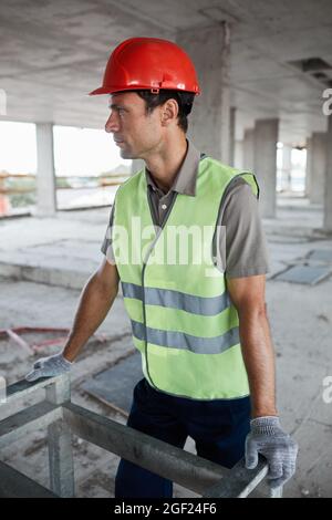 Vertical portrait of male construction worker wearing hardhat and reflective jacket at construction site and looking away Stock Photo