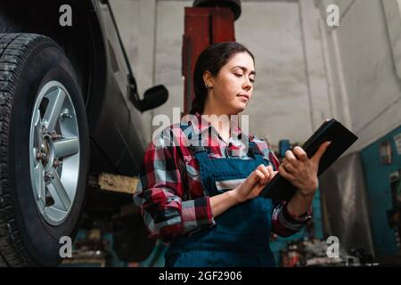 Portrait of a young Caucasian female mechanic, in uniform, using a tablet in her hands. The wheel of the car on the right. Bottom view. Stock Photo