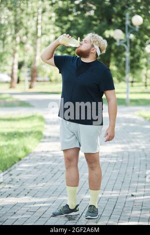 Overweight man standing outdoors and drinking water from the bottle after training in the park Stock Photo