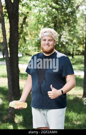 Portrait of happy overweight man with bottle of water smiling at camera and showing thumb up after training in the park Stock Photo