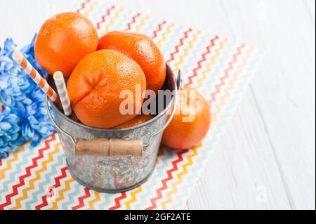 Close up of fresh clementines in a tin bucket with straws on a wooden table. Chevron pattern napkin and blue flowers Stock Photo