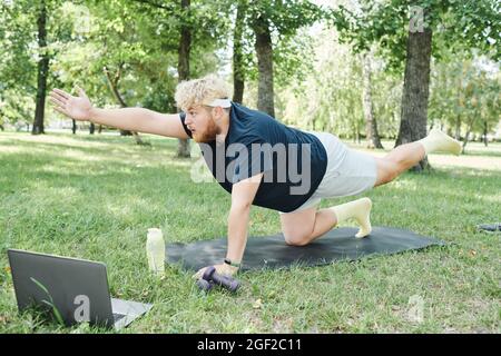 Overweight man exercising with dumbbells on exercise mat in the park and watching sports training online on laptop Stock Photo