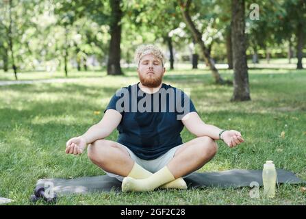 Overweight man sitting on the grass with eyes closed and meditating after sports training in the park Stock Photo