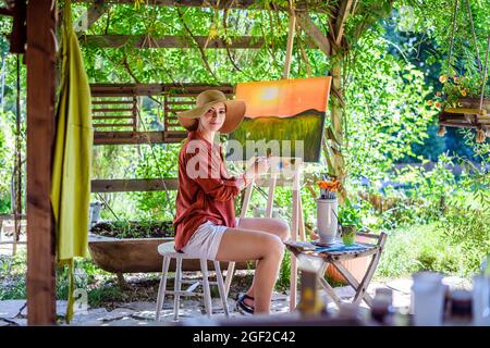 Young female artist working on her art canvas painting outdoors in her garden. Mindfulness, art therapy, creativity and creative hobbies concept. Stock Photo
