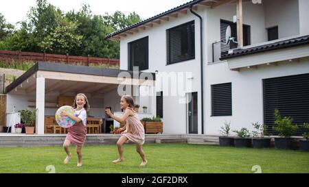 Two small girls sisters playing with a ball outdoors in the backyard, running. Stock Photo
