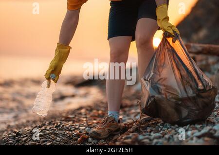 Earth day. Cleanup garbage on the ocean coast. A volunteer with polyethielene bag picking up a plastic bottle on the beach. Close-up. Sunset in the ba Stock Photo