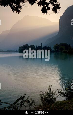 Sundown in September over an alpine lake in northern Italy, the Lago di Toblino in Trentino-Alto Adige.  A medieval castle, the Castel Toblino, is in silhouette as beams from the sinking sun light rugged landscape between the Dolomite peaks of Monte Casale and Monte Garzole or Piccolo Dain. Stock Photo