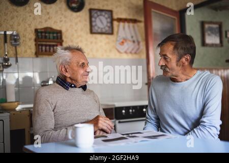Portrait of man with elderly father sitting at the table indoors at home, talking. Stock Photo