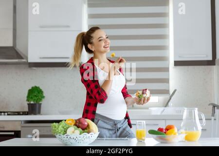 Pregnant blond young woman in red checkered shirt at kitchen eating a fruit salad, smiling, looking away, in front of her bowls with fresh vegetables, juice and fruits. Healthy eating concept. Stock Photo