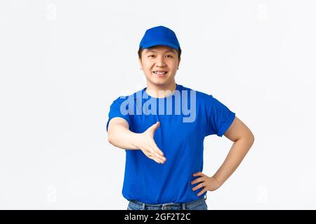 Friendly cheerful asian courier in blue uniform smiling confident, extend hand for handshake, greeting customer, making deal. Delivery man saying Stock Photo