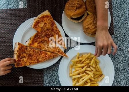 children's hands to take fast food on the table. Pizza, hamburger, fries. Stock Photo