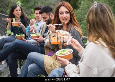 young people gathering at the park. Reunion of best friends eating together salads in take away cardboard bowls sitting outdoors and having fun. Stock Photo