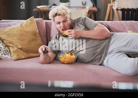 Lazy overweight man lying on the sofa eating chips and watching TV at home Stock Photo