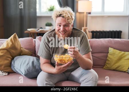 Overweight man sitting on sofa with bowl of chips eating them and laughing while watching TV at home Stock Photo