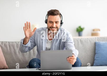 Distant communication concept. Happy millennial man in headset waving hand at laptop camera, sitting on sofa Stock Photo