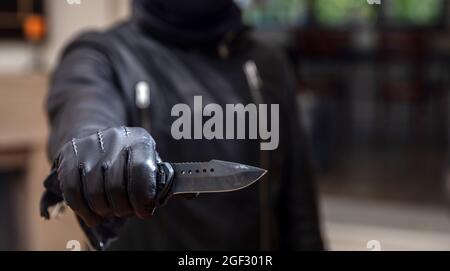 Robber threaten with a dagger, blur indoor background, closeup view. Burglar  holding a knife in gloved hand, armed robbery concept. Stock Photo