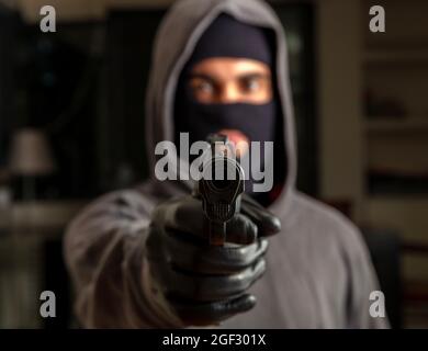 Terrorist hooded in balaclava, holding a gun in gloved hand. Man aiming with a pistol, dark building indoor background, Killer, armed robbery concept. Stock Photo