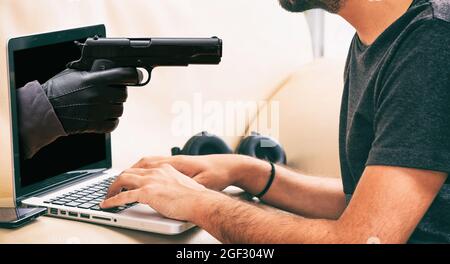 Gun and laptop. Gloved hand holding pistol aiming at young man working with computer. Cyber crime, hacker and stealing information concept. Stock Photo