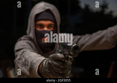 Killer hooded with balaclava, holding a gun in gloved hand, dark outdoors background, armed robbery concept. Stock Photo