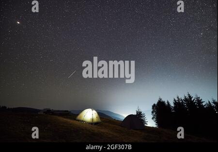 Night camping in the mountains. Two tents, one of which illuminated, set up on mountain meadow. Panoramic view of evening sky with multiple stars. Stock Photo