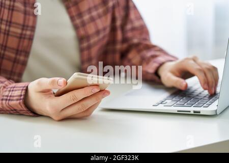 Faceless young female browsing smartphone while typing on laptop. Millennial data privacy concept Stock Photo