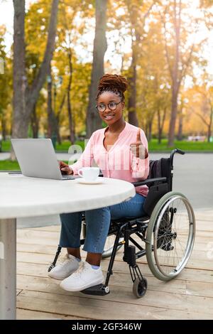 Disabled black lady using laptop, working online, showing thumb up at autumn park. Remote job for handicapped people Stock Photo