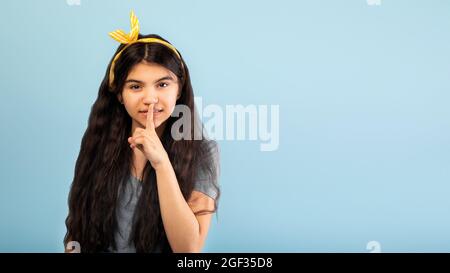 Mysterious Indian teenage girl showing shhh sign at camera, putting finger on lips, gesturing hush over blue background Stock Photo