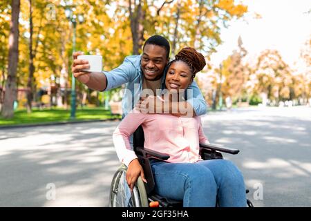 Cheerful black guy hugging his disabled girlfriend in wheelchair, making selfie on smartphone at autumn park Stock Photo