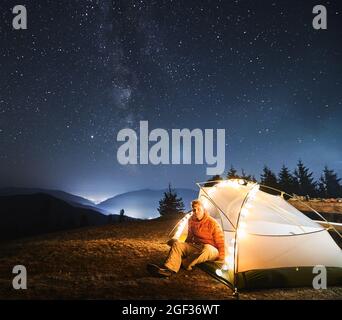 Young traveler sitting in his white tent, lit by light bulbs, on grass under open starry sky with Milky way. Night mountain camping against backdrop of mountain hills and light of city between them. Stock Photo