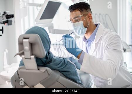 Middle-Eastern Male Dentist In Medical Mask And Face Shield Checking Patient's Teeth Stock Photo