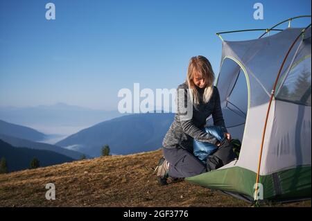 Blonde female traveler packing her sleeping bag in protective bag after night's rest, sitting down at the edge of tent. Vacation in the mountains in tourist campsite. Concept of travelling and camping Stock Photo
