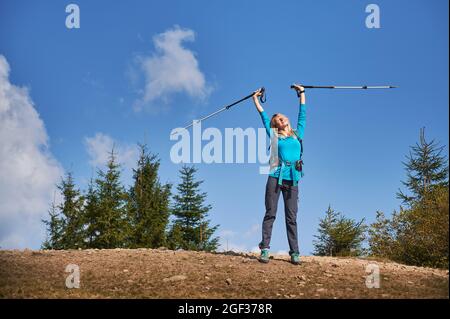 Pleasured female hiker with high up arms in which she holding sticks for walking, closed her eyes and basking in sun, standing on rocky surface in fresh air against blue sky. Stock Photo