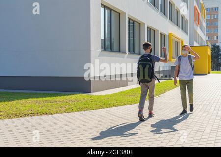 Children waving hand, greeting friend on street. Kids with face masks and backpacks meet near school building outdoors. Back to school, friendship Stock Photo