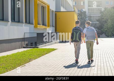 Children with face masks and backpacks meet near school building and walk to the school. Back to school, friendship Stock Photo
