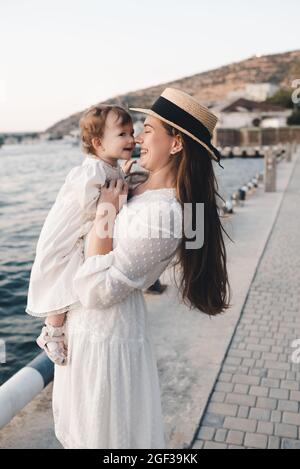 Happy cheerful baby girl sitting on mother hands wear stylish white dresse and straw hat relax over sea shore at quayside outdoors close up. Looking a Stock Photo