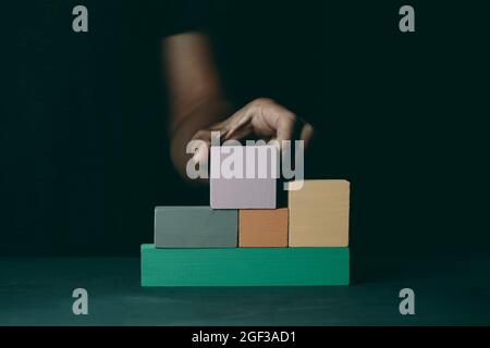 a caucasian man builds a structure with some wooden toy blocks of different colors, on a dark gray surface Stock Photo