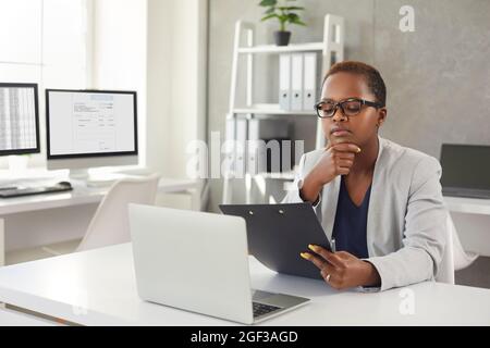 Black woman sitting at her desk in office, reading business documents and thinking Stock Photo