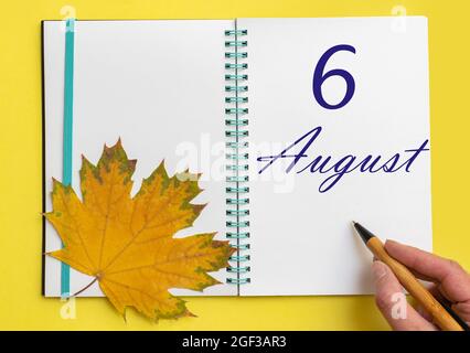 6th day of august. Hand writing the date 6 august in an open notebook with a beautiful natural maple leaf on a yellow background. Summer month, day of Stock Photo