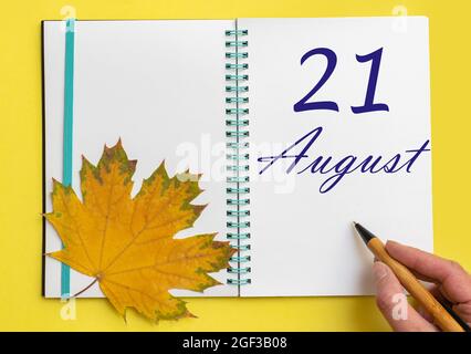 21st day of august. Hand writing the date 21 august in an open notebook with a beautiful natural maple leaf on a yellow background. Summer month, day Stock Photo