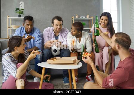 Group of happy diverse friends eating pizza, drinking beer and discussing soccer match Stock Photo