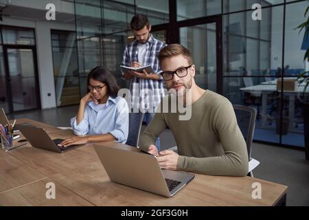 Young caucasian man, male office worker using laptop, sitting at office desk and looking at camera. Group of young multicultural business people Stock Photo