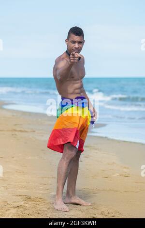 Man with a colourful gay pride flag and pointing to camera while standing on the beach. Stock Photo