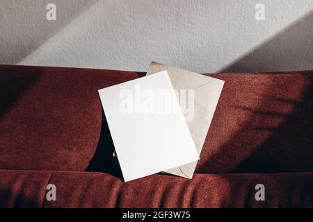 Blank greeting card mockup, envelope on rusty velvet sofa in sunlight. Empty invitation over white wall with shadows. Detail of elegant interior Stock Photo
