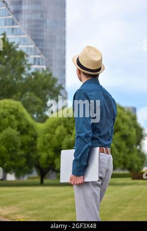 A man with a laptop in his hand stands and looks towards the office building. Young student, programmer and businessman with computer in hand Stock Photo