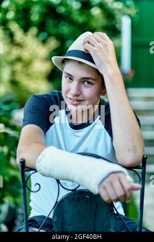 Portrait of young caucasian boy with a broken and cast arm wearing a hat and sitting in a chair outdoor in a garden. Lifestyle concept. Stock Photo