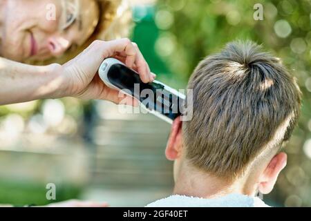 Portrait of a mother cutting young caucasian boy's hair outside in a garden. Lifestyle concept. Stock Photo