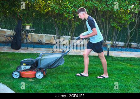 Portrait of a young caucasian boy passing the mower through  in the garden in a country house. Lifestyle concept. Stock Photo