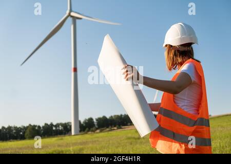 An engineer of wind turbines holding a paper project and looking at windmill in the field. Green eco electricity generation and renewable energy.  Stock Photo