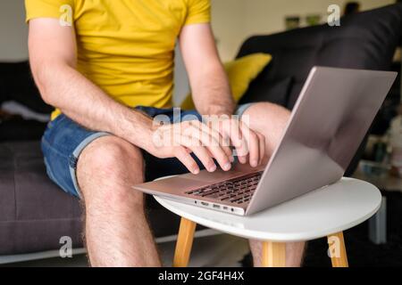 Man in the yellow T shirt typing on the laptop sitting in the modern living room. Freelancer working at home Stock Photo