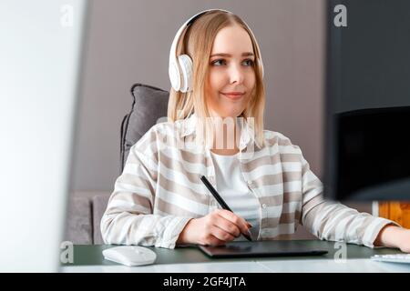Young woman freelancer designer use graphic tablet while working. Digital artist Woman in headphones works using pc computer At home office. Remote Stock Photo
